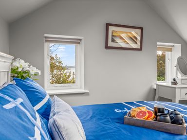 A bedroom with a bed with blue bedding a tray of breakfast on the bed two windows and a vanity table at Seascape in Kingsand
