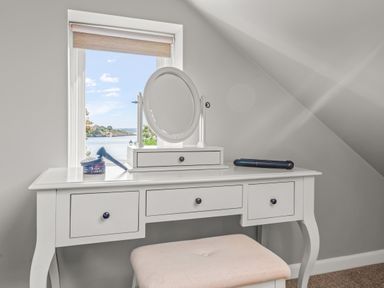 A white dressing table with a round mirror and a stool in front of a window with a view of water at Seascape Kingsand