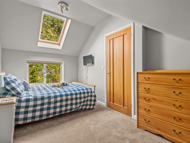 A bedroom with a blue checkered bedspread wooden dresser wooden door and windows showing greenery at Seascape in Kingsand