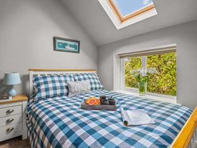 A bedroom with a bed and blue checkered bedding and a tray with breakfast and a magazine on the bed near a window with flowers and a skylight at Seascape in Kingsand