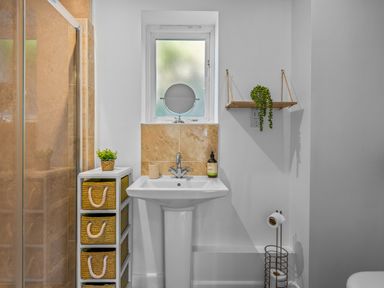A bathroom with a pedestal sink baskets on a shelf and a window above the sink at Seascape in Kingsand