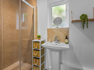 A bathroom with a white sink pedestal a mirror on the windowsill a shower with glass doors a storage unit with baskets a small plant on the unit and a hanging shelf with a plant at Seascape in Kingsand