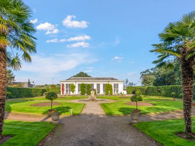 A garden with trimmed lawns, small trees in pots, a central fountain, and a glass building at Seascape in Kingsand