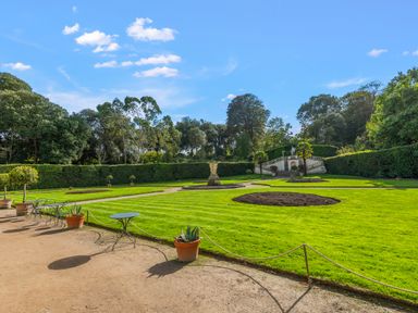 A garden with green lawns, potted plants, metal tables and chairs, a stone statue on a pedestal, and trees in the background at Seascape in Kingsand