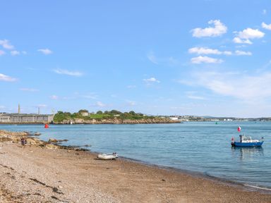 A beach with pebbles and rocks a small boat on water and an island with buildings across the bay at Seascape in Kingsand