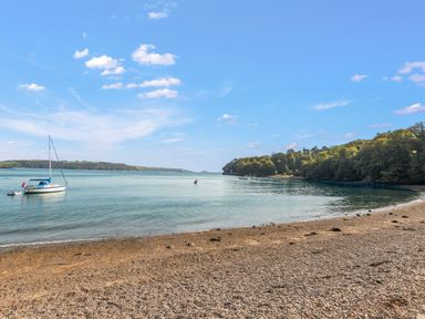 A pebble beach with boats on calm water and trees on the shoreline at Seascape in Kingsand