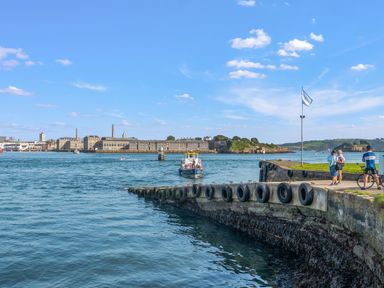 A waterfront with a stone pier and tires lining the edge with people walking and a boat on the water at Seascape in Kingsand