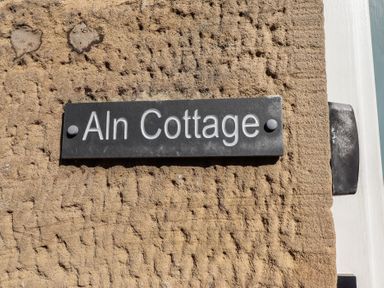 A black sign with white letters reading Aln Cottage mounted on a rough beige wall at Aln Cottage in Alnmouth