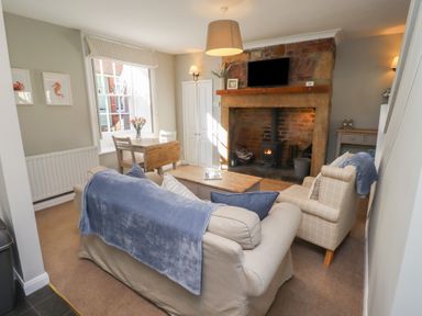 A living room with a sofa and armchair facing a fireplace with a wood stove and a wall-mounted TV at Aln Cottage (Alnmouth) in Alnmouth