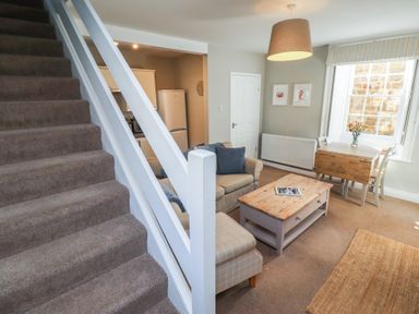 A living area with carpeted stairs a beige sofa a coffee table and a wooden dining table by a window at Aln Cottage Alnmouth