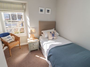 A bedroom with a single bed blue blanket bedside table lamp wicker chair and window at Aln Cottage Alnmouth