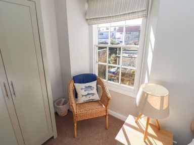 A corner of a room with a wicker chair with a fish cushion next to a window a lamp on a wooden table and a green wardrobe at Aln Cottage Alnmouth