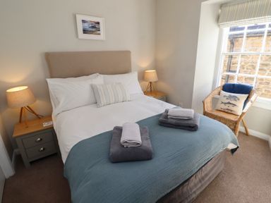 A bedroom with a bed with folded towels bedside tables with lamps a wicker chair and a window at Aln Cottage Alnmouth