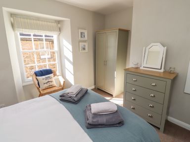 A bedroom with a bed covered in folded towels a wicker chair by a window a wardrobe and a chest of drawers with a mirror at Aln Cottage Alnmouth