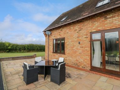 A patio with a table and chairs outside a brick house with glass doors at Little Acre in Church Lench near Harvington