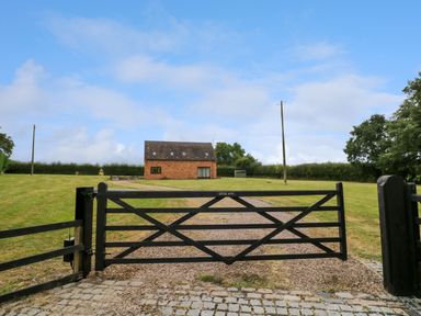 A black gate opens to a gravel driveway leading to a brick house with multiple windows and a grassy lawn at Little Acre in Church Lench near Harvington