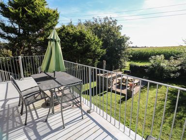 A balcony with a table and four chairs and a folded green umbrella overlooking a garden with a picnic table and barbecue at Tremornah in Lamorna