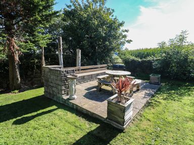 A patio with a round wooden picnic table and benches a stone wall bench a barbecue and planter boxes in a garden at Tremornah in Lamorna