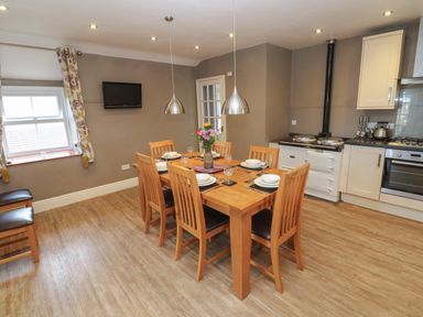 A dining area with wooden table and six chairs a kitchen with appliances and a window with curtains at Tremornah in Lamorna