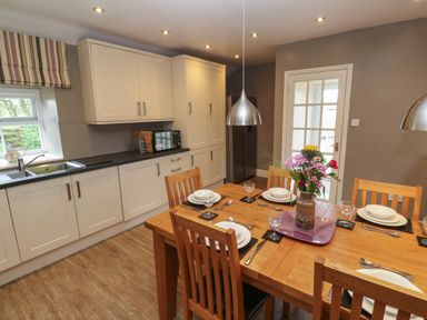 A kitchen with wooden dining table set for six with flowers in a vase and white cabinets at Tremornah in Lamorna