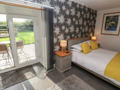 A bedroom with a bed yellow pillows bedside tables lamps a door leading to a garden and patterned wallpaper at Tremornah in Lamorna