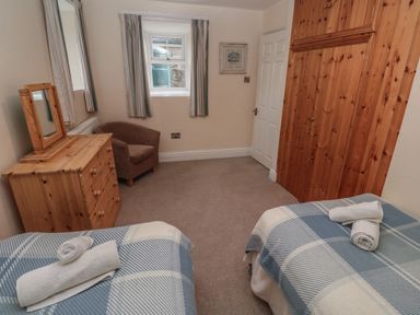 A bedroom with two single beds covered with checkered blankets and towels on them a wooden chest of drawers with a mirror a brown armchair and wooden built-in wardrobes at Tremornah in Lamorna