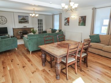 A dining area with a wooden table and four chairs and a living area with green and beige sofas a fireplace and TV at Tremornah in Lamorna