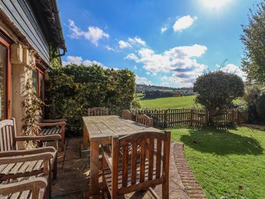An outdoor patio with wooden table and chairs overlooking a grassy garden and field at Highgrove Barn in Painswick