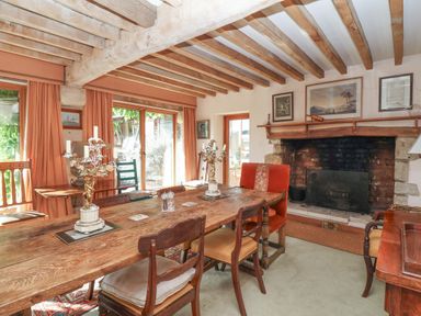 A dining room with a wooden table and chairs near a fireplace and windows with curtains at Highgrove Barn in Painswick