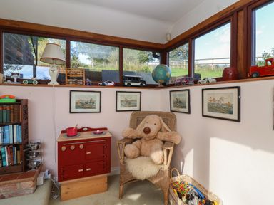 A corner room with a wicker chair holding a large stuffed dog toy a red toy stove a basket of toys books on a shelf and framed pictures on the walls at Highgrove Barn in Painswick