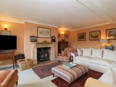 A living room with a fireplace a sofa armchairs a patterned ottoman a television and framed pictures at Highgrove Barn in Painswick