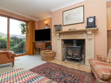 A living room with a fireplace a television and sliding glass doors showing greenery at Highgrove Barn in Painswick