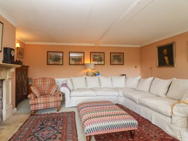 A living room with a large white sectional sofa a patterned ottoman a plaid armchair framed pictures on the wall and a fireplace at Highgrove Barn in Painswick