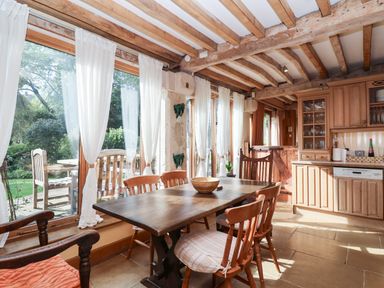 A dining area with wooden table and chairs next to large windows with white curtains and a kitchen area with wooden cabinets at Highgrove Barn in Painswick