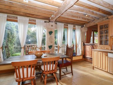 A dining area with wooden table and chairs near large windows with white curtains and a wooden cabinet at Highgrove Barn in Painswick