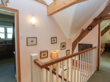 A stairwell with framed pictures on the walls a lit table lamp and visible doorways leading to adjacent rooms at Highgrove Barn in Painswick
