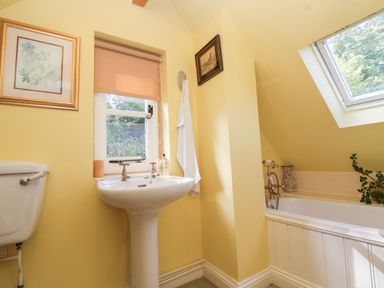 A bathroom with a pedestal sink, toilet, bathtub with a window above and framed pictures on yellow walls at Highgrove Barn in Painswick
