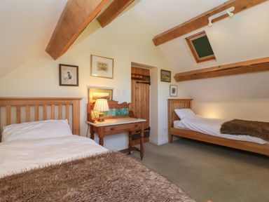 A bedroom with two single wooden beds a small wooden desk with a lamp and framed pictures on cream walls at Highgrove Barn in Painswick