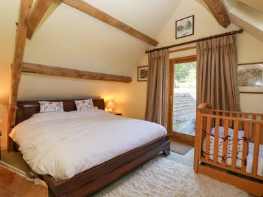 A bedroom with a large bed a wooden crib a door with curtains and exposed wooden beams at Highgrove Barn in Painswick