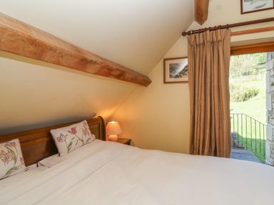 A bedroom with a wooden bed frame floral pillows a bedside table with a lamp a curtain and a door leading outside at Highgrove Barn in Painswick