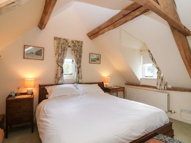 A bedroom with a large bed two bedside tables with lamps two small windows with floral curtains and wooden beams at Highgrove Barn in Painswick