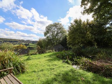 A grassy garden area with a wooden bench a small pond trees and a fence with hills in the background at Highgrove Barn in Painswick