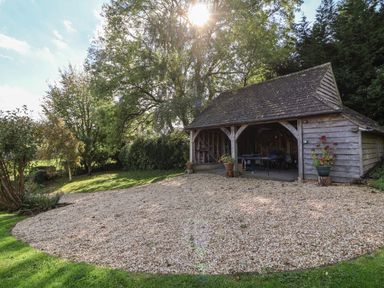 A wooden open garage with a gravel driveway and trees around at Highgrove Barn in Painswick