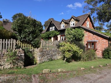 A brick house with dormer windows surrounded by bushes and trees with a stone wall and wooden fence in front at Highgrove Barn in Painswick