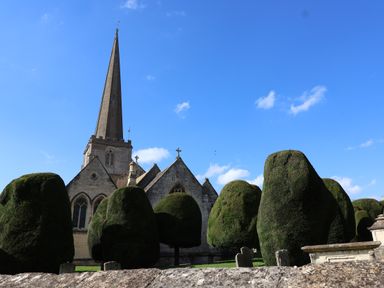 A stone church with a tall spire behind trimmed shrubs and gravestones at Highgrove Barn in Painswick