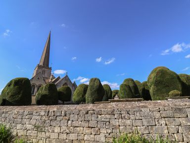 A stone church with a tall spire behind a stone wall and shaped green bushes under a blue sky