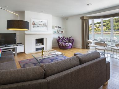 A living room with a gray sofa, glass coffee table, fireplace, wall art, TV, purple armchair, dining table, and large windows overlooking a waterfront at 21 Dart Marina in Dartmouth