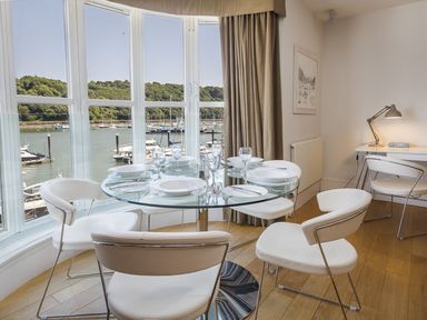 A dining area with a round glass table set with plates and glasses near large windows showing a marina at 21 Dart Marina in Dartmouth