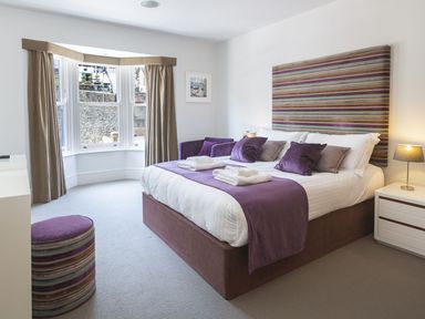 A bedroom with a bed purple cushions and a purple throw near a bay window with beige curtains at 21 Dart Marina in Dartmouth