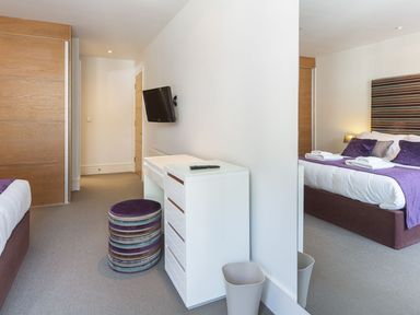 A bedroom with a double bed with purple bedding a wall-mounted television a white chest of drawers a round stool and a large mirror at 21 Dart Marina Dartmouth